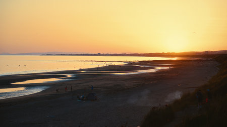 Sunset over Irvine Beach, Scotland, on 12 July 2025. Warm evening light reflecting on calm sea waves and sandy shore with silhouettes of people walking.のeditorial素材