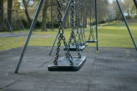 Close-up view of empty swings hanging in a quiet park on a calm day. The image conveys a nostalgic and peaceful mood, surrounded by green grass and trees. Linlithgow, Scotlandの写真素材