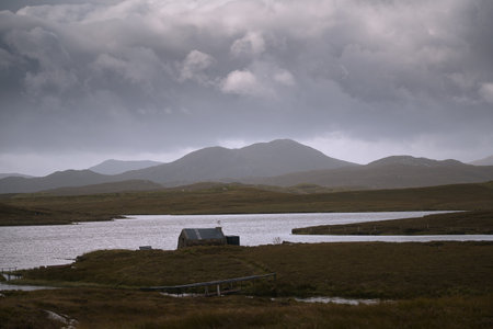 A small cabin by a quiet lake on the Isle of Lewis, Scotland. Moody clouds and distant hills create a peaceful yet dramatic atmosphere in the Outer Hebrides landscape.の写真素材