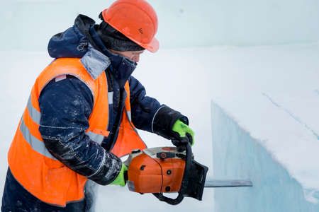 Worker fitter in a protective helmet and reflective vest sawing an ice plate with a chainsaw