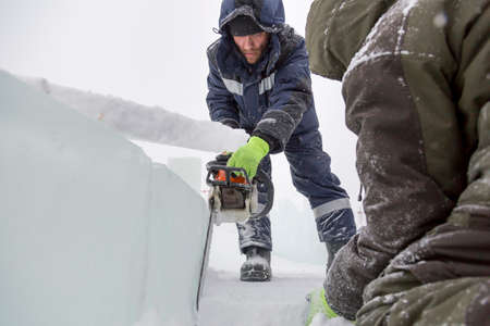 Workman assembler in a blue jacket with a chainsaw in his hands cuts ice block