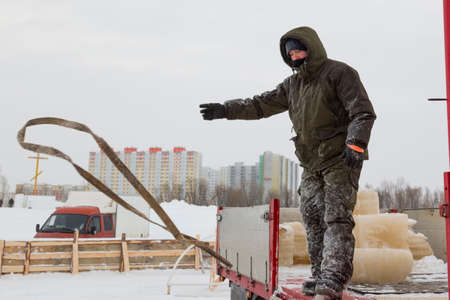 Worker in a green winter suit with a hood on his head with slings in his hand