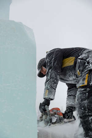 A sculptor cuts an ice figure out of a block of ice with a chainsaw against the backdrop of a Christmas tree.の写真素材