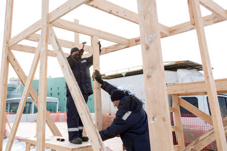 Builder with a hammer in his hand in a blue jacket with a hood on the assembly of a wooden structure