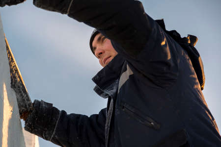 A worker fitter in a blue jacket with a hood on his head drives an ice plate with a chainsaw