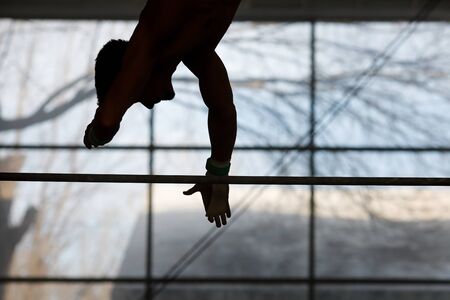 Silhouette of a young male gymnast training at the Horizontal Bar in a gym.