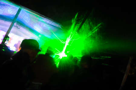 Trippy night image with people dancing outdoors in a forest music festival with laser beams in the...