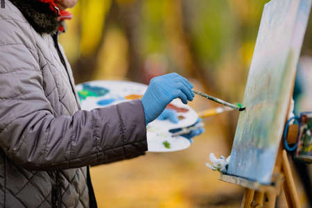 Shallow depth of field (selective focus) image with the hand of a female painter and her color palette while painting a landscape on a sunny autumn day.の写真素材