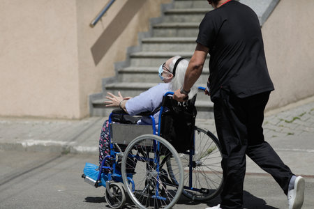 Bucharest, Romania - May 9, 2022: Orderly man pushes a senior male patient in a wheelchair on the concrete in the court of a Romanian hospital.のeditorial素材