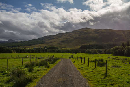 lonely road in a green field surrounded by mountainsの写真素材