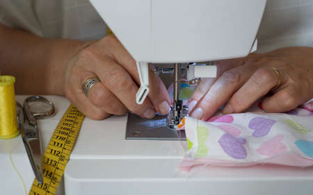 Detail of seamstress hands working at white sewing machine. She is using a hearts fabric, yellow thread, scissors, and yellow sewing meterの写真素材