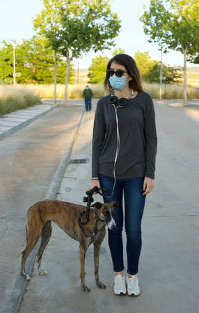 Young woman wearing face mask and sunglasses walking with a galgo dog on a street during coronavirus outbreak. Global COVID-19 pandemic concept image. New normal lifestyle for prevention of spreading COVID-19の写真素材