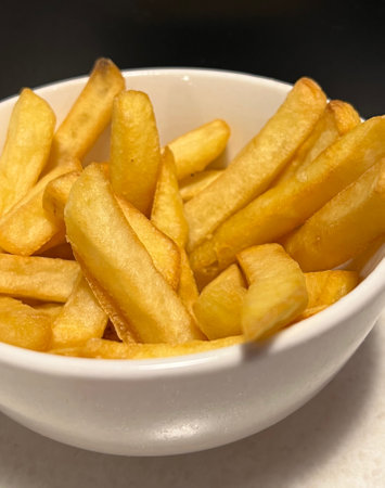 Close-up of a portion of chips in a white bowl on a black background. French fries, fast food,...