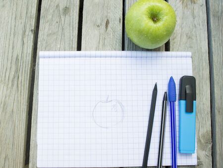 squared notebook with a drawing of an apple, a pen, a pencil and a blue marker that lies on top, in the background is a green apple on a wooden surfaceの写真素材