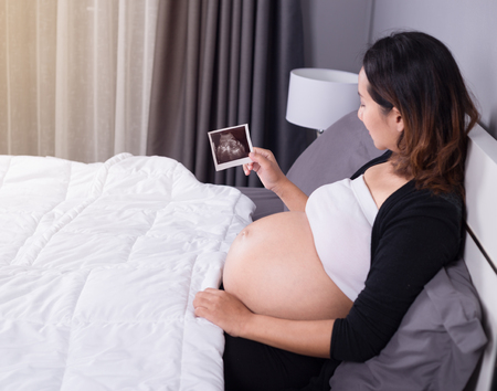 pregnant woman with ultrasound image of baby sitting on bed in the bedroom at homeの写真素材