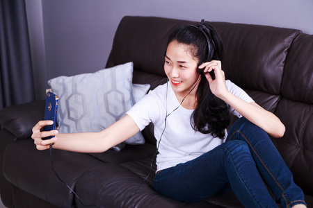woman enjoying music in headphones, making selfie photo with cellphone while lying on sofa at homeの写真素材