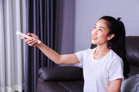 young woman holding a remote control air conditioner on sofa at homeの写真素材