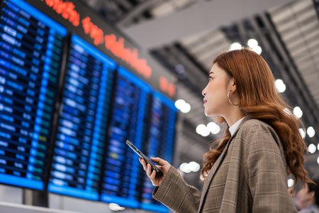 beautiful woman using smartphone with flight information board at airportの写真素材