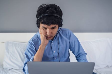 stressed business man wear headset video conference calling on laptop computer on a bed, work from...