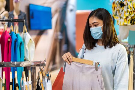 young woman choosing clothes at shopping mall and her wearing medical mask for prevention from...
