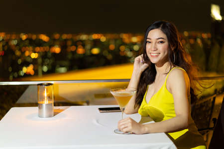 beautiful woman in evening dress holding cocktail at restaurant over night city backgroundの写真素材