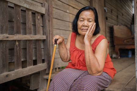Thai elderly woman in round-necked sleeveless collar with headache and worried stressed face in old wooden homeの写真素材