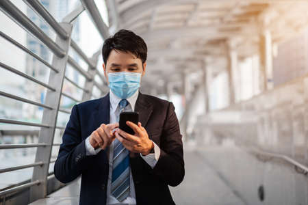 business man using a smartphone and wearing medical mask during coronavirus (covid-19) pandemic in the cityの写真素材