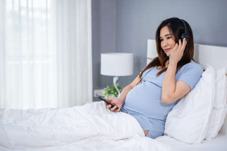 pregnant woman using smartphone to listening music through headphones on a bedの写真素材