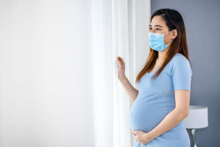 pregnant woman in medical mask and looking through window in a bedroom, stay alone for protect...