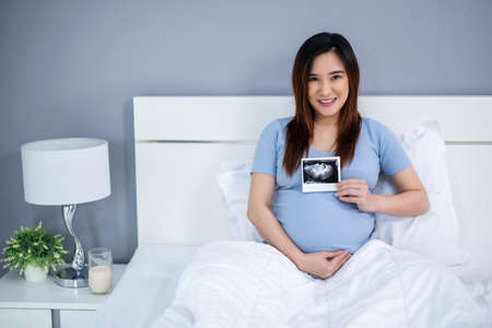 happy pregnant woman holding ultrasound scan photo on a bedの写真素材