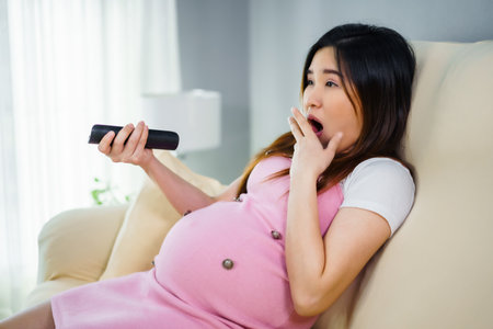 shocked pregnant woman watching tv with remote control on sofa in the living roomの写真素材