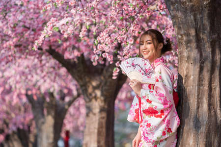 woman in yukata (kimono dress) holding folding fan and looking sakura flower or cherry blossom blooming in the gardenの写真素材