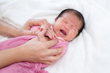 crying newborn baby wrap in blanket and parent hands on a bedの写真素材