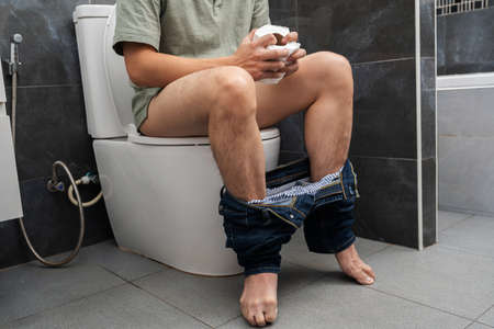 man with stomach issues sitting in toilet bowl and holding tissue roll in the bathroom