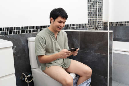 happy man using smartphone and sitting in toilet bowl in the bathroom at homeの写真素材