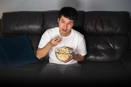 young man watching TV and eating popcorn on sofa at nightの写真素材