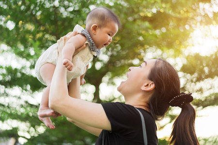 mother holding baby newborn in her arms lifting up on a green nature bokeh background with sunlightの写真素材