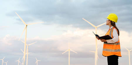 young female engineer using walkie talkie and reading clipboard to checking system against wind turbine farmの写真素材