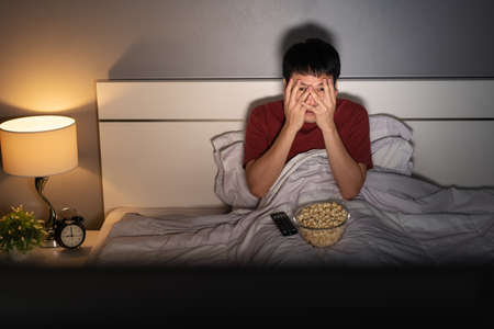scared young man is watching horror movie TV on a bed at night