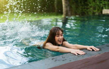 young woman hold on to the edge of the swimming pool and splashing water with her feetの写真素材