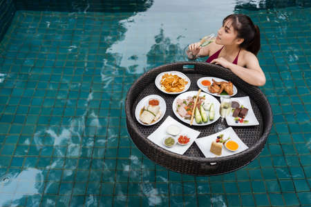 cheerful young woman enjoying with floating food and drinking champagne glass in swimming poolの写真素材
