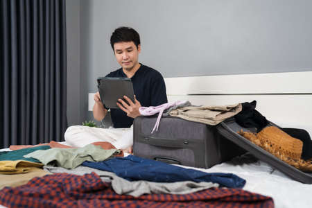young man writing tablet and packing clothes into suitcase on a bed at home, planning travel holiday.の写真素材