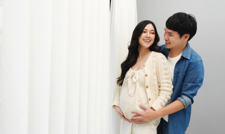 married couple is expecting baby. man embracing and talking with his pregnant wife while looking view through the window at homeの写真素材