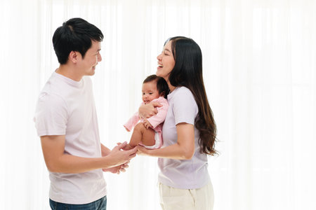 cheerful father and mother holding and playing with newborn baby on white window background. happy family conceptの写真素材