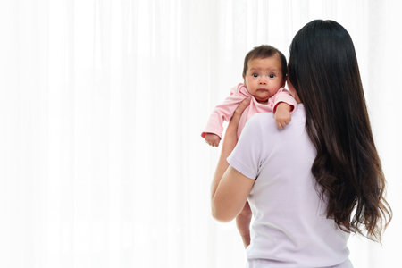 mother holding newborn baby burping after feeding milk on white window backgroundの写真素材