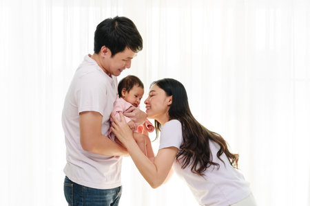 cheerful father and mother holding and playing with newborn baby on white window background. happy family conceptの写真素材