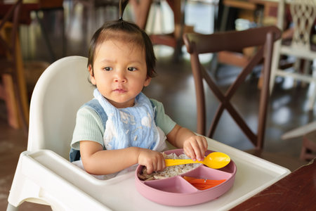toddler girl sitting on highchair eating food and vegetable in the restaurantの写真素材
