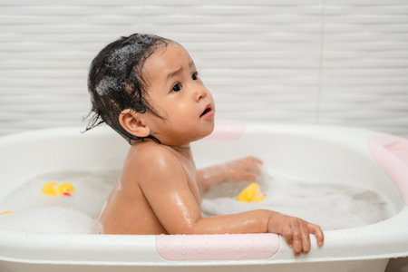 happy infant baby take a bath and playing with foam bubbles in bathtub at homeの写真素材