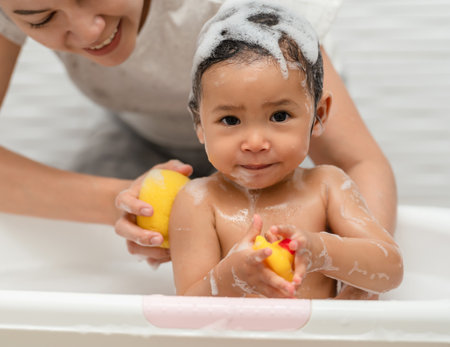 mother bathing her cheerful infant baby with a soft sponge with foam bubbles in a bathtubの写真素材