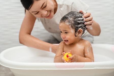 mother bathing and washing her infant baby hair with shampoo while playing a rubber duck toy in a...